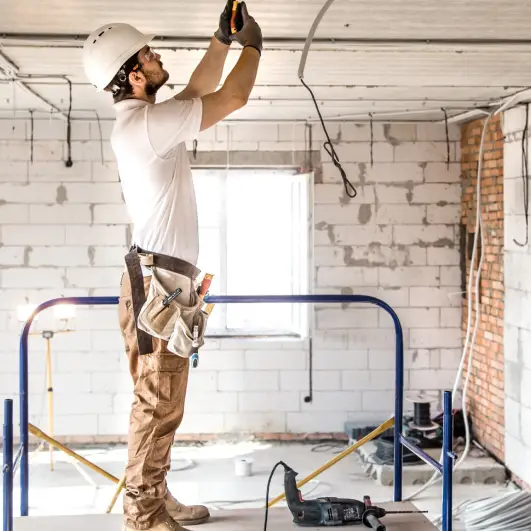 A worker in a hard hat stands on a scaffold, using a tool to install ceiling fixtures in a partially constructed room.