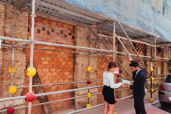 A man and woman discuss plans in front of a building under renovation, surrounded by scaffolding and construction materials.